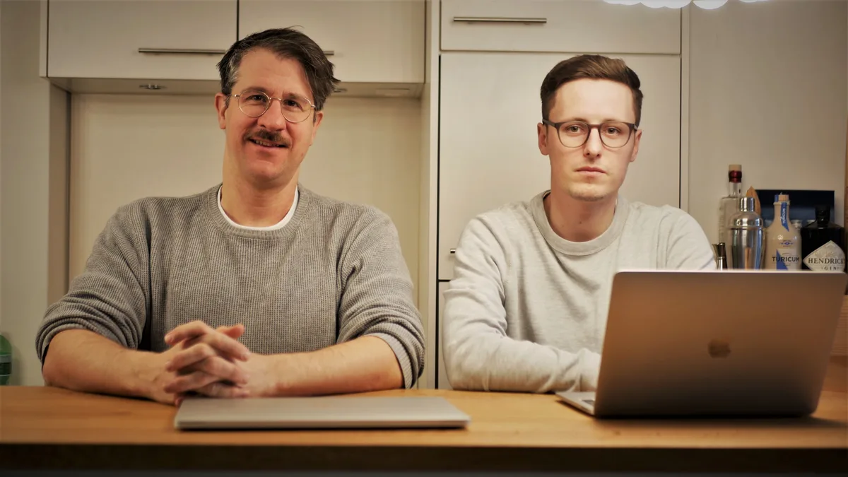 Man sieht zwei Männer an einer Bar stehen, hinter ihnen steht ein Kühlschrank, Wandschränke und ein Regal mit Schnapsflaschen.